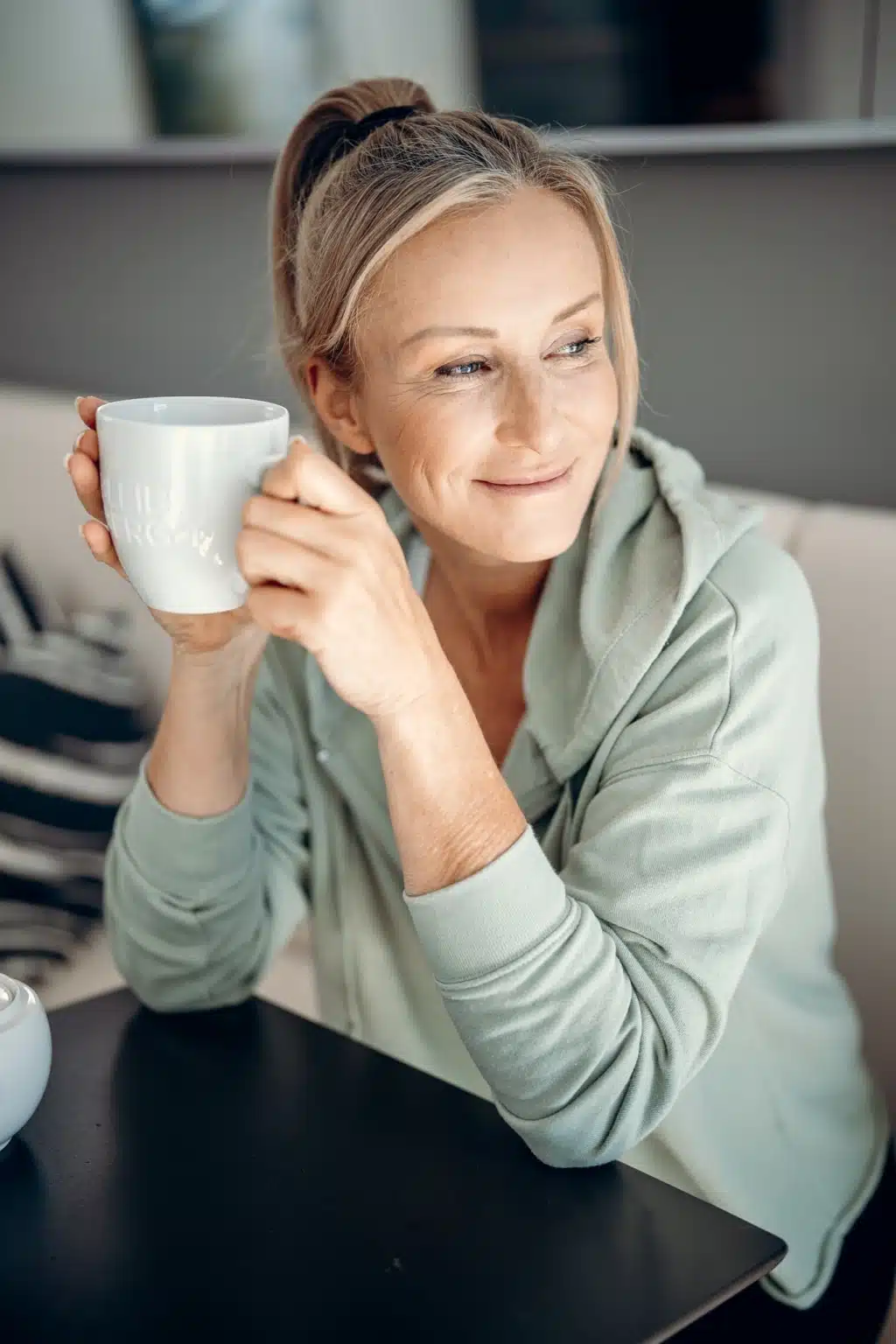 blonde Frau sitzt an einem Tisch im BLEIB BERG F.X. Mayr Retreat und hält lächelnd eine Tasse Tee